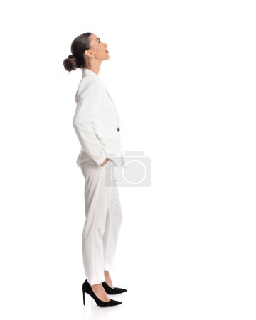 Téléchargez les photos : Vue latérale de femme d'affaires attrayante tenant la main dans les poches et regardant vers le haut en face de fond blanc - en image libre de droit