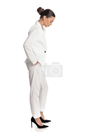 Téléchargez les photos : Vue latérale de l'élégante femme de mode en costume blanc tenant les mains dans les poches, regardant vers le bas et posant sur fond blanc - en image libre de droit