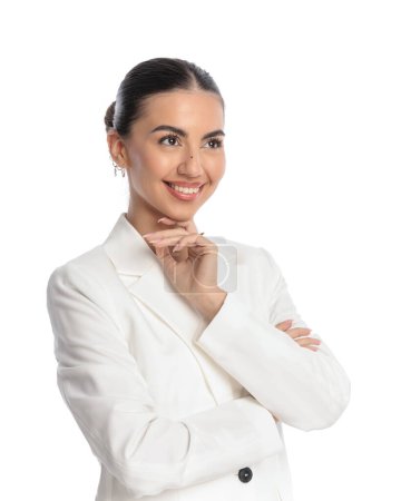 Téléchargez les photos : Portrait de belle jeune femme d'affaires en costume blanc regardant loin, touchant le menton et la pensée, souriant et croisant les bras sur fond blanc - en image libre de droit