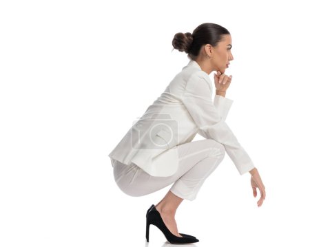 Foto de Imagen de cuerpo completo de mujer joven elegante en traje blanco tocando la barbilla y pensando mientras sostiene los codos en las rodillas y agacharse sobre fondo blanco - Imagen libre de derechos