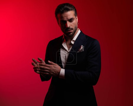 Foto de Sensual hombre de negocios elegante en traje con camisa de cuello abierto frotando las palmas y siendo seductor delante de fondo rojo - Imagen libre de derechos