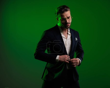 Photo for Portrait of attractive elegant man with glasses looking away and buttoning suit in front of green background in studio - Royalty Free Image
