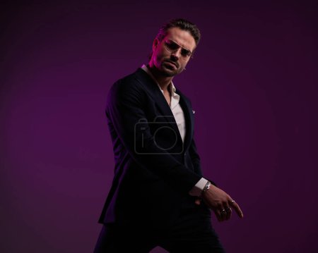 Photo for High class businessman with glasses holding hand in pocket and posing with arms in cool pose in front of purple background - Royalty Free Image
