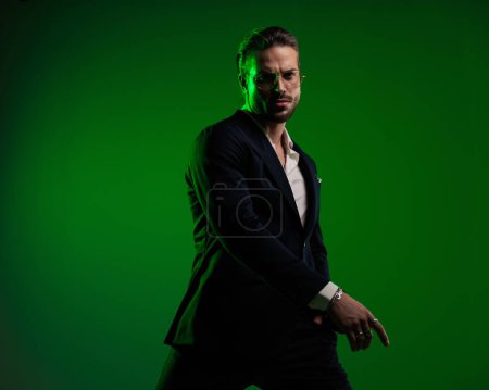 Téléchargez les photos : Sexy homme élégant avec des lunettes tenant les bras dans la pose de mode avec la main dans les poches en face de fond vert - en image libre de droit