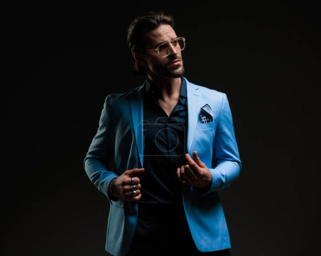 Foto de Retrato de hombre barbudo sexy con gafas mirando a un lado mientras se ajusta traje azul y posando de una manera segura delante de fondo gris oscuro - Imagen libre de derechos