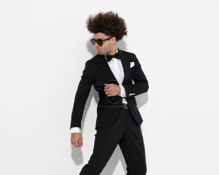 Photo for Attractive fashion man with curly hair and sunglasses looking to side and dancing in front of grey background - Royalty Free Image