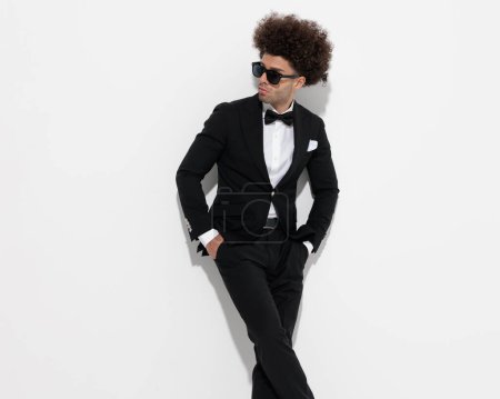 Photo for Confident man with big curly hair holding hands in pockets and looking to side while wearing high class tuxedo in front of grey background - Royalty Free Image