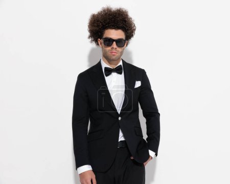 Téléchargez les photos : Portrait d'un homme d'affaires haut de gamme en smoking noir avec des lunettes de soleil tenant la main dans les poches et posant de manière confiante sur fond gris - en image libre de droit