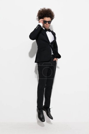 Photo for Cool high class man in black tuxedo holding hand in pocket, fixing sunglasses and jumping in the air in front of grey background - Royalty Free Image