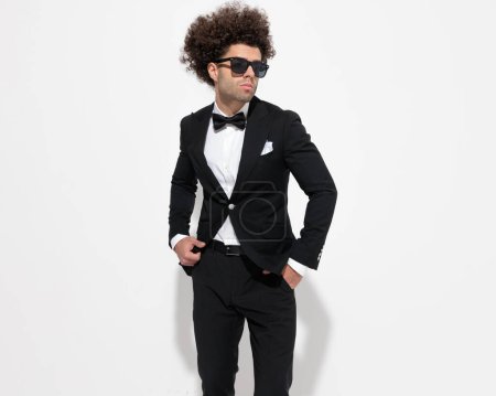 Photo for Elegant young groom in black tuxedo with sunglasses holding hands in pockets and looking to side in front of grey background - Royalty Free Image