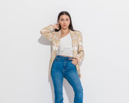 Photo for Sexy smart casual woman in paisley print jacket holding hand in jeans pockets while adjusting hair and posing in front of grey background - Royalty Free Image