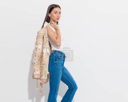 Photo for Side view of cool hippie girl holding paisley print jacket over shoulder and posing with hand in pocket in front of grey background - Royalty Free Image