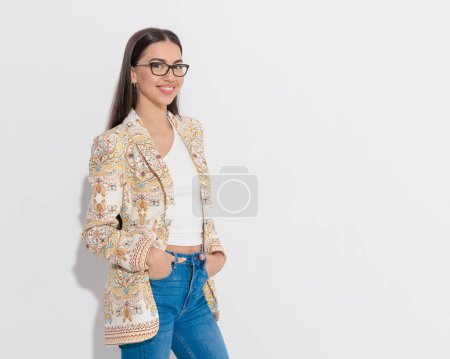 Photo for Sexy hippie girl with paisley print jacket and glasses holding hands in pockets and smiling in front of grey background - Royalty Free Image