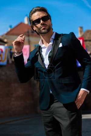 Téléchargez les photos : Homme d'affaires de mode avec chemise à col ouvert et lunettes de soleil tenant la main dans les poches et regardant vers le haut en plein air devant la ville médiévale - en image libre de droit