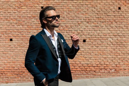 Téléchargez les photos : Vue de côté image de jeune homme d'affaires sexy en costume avec des lunettes de soleil regardant vers le côté et marchant à l'extérieur en face de fond de la ville médiévale - en image libre de droit