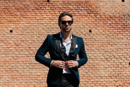 Foto de Retrato de elegante hombre de negocios barbudo con gafas de sol fijando traje y caminando al aire libre en una ciudad vieja de Transilvania - Imagen libre de derechos