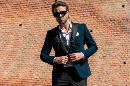 Téléchargez les photos : Portrait de sexy élégant homme d'affaires avec des lunettes de soleil boutonnage costume bleu marine et posant en plein air dans une vieille ville de Roumanie - en image libre de droit