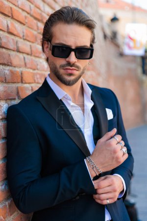 Téléchargez les photos : Portrait d'un homme de mode confiant avec des lunettes de soleil arrangeant les manches de chemise et posant en plein air dans une vieille ville de Roumanie - en image libre de droit