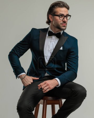 Photo for Sexy young man with glasses looking to side and sitting with arms on thighs in front of grey background - Royalty Free Image