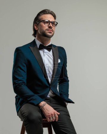 Foto de Pensativo hombre de negocios elegante con gafas en esmoquin mirando hacia arriba, pensando y sentado con la mano en el bolsillo delante de fondo gris oscuro - Imagen libre de derechos