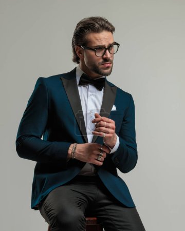 Photo for Portrait of sexy elegant man with glasses in tuxedo looking to side and sitting while adjusting shirt sleeves on grey background - Royalty Free Image