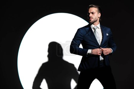 Téléchargez les photos : Homme élégant confiant regardant de côté et fermeture costume bleu marine et regardant de côté tout en posant devant projecteur studio sur fond blanc - en image libre de droit