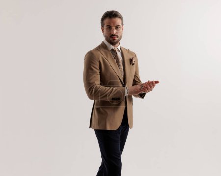 Foto de Hombre de negocios de moda cool en traje marrón frotando las palmas y moviéndose delante de fondo gris - Imagen libre de derechos
