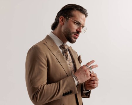 Photo for Portrait of side view of sexy bearded man with glasses looking to side and arranging rings while confidently posing on grey background - Royalty Free Image
