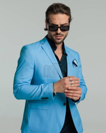Téléchargez les photos : Portrait de sexy élégant homme en costume bleu avec des lunettes de soleil regardant vers le bas tout en fixant des anneaux devant fond gris - en image libre de droit