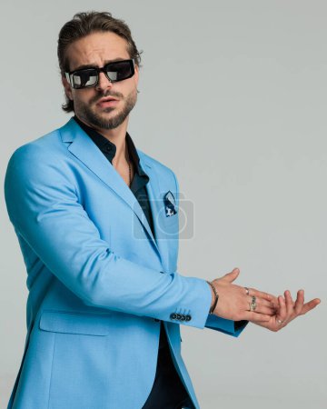 Foto de Retrato de hombre de negocios de moda fresco en traje azul con gafas de sol frotando las palmas a un lado delante del fondo gris - Imagen libre de derechos