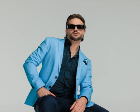 Téléchargez les photos : Cool homme de mode en costume bleu avec des lunettes de soleil assis et posant avec les bras sur les cuisses en face de fond gris - en image libre de droit