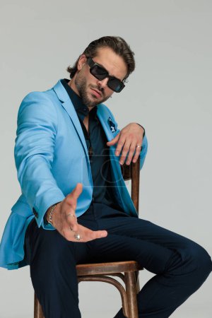 Téléchargez les photos : Élégant homme d'affaires de la mode avec des lunettes de soleil assis et faisant geste de la main, pointant la main vers l'avant sur fond gris - en image libre de droit