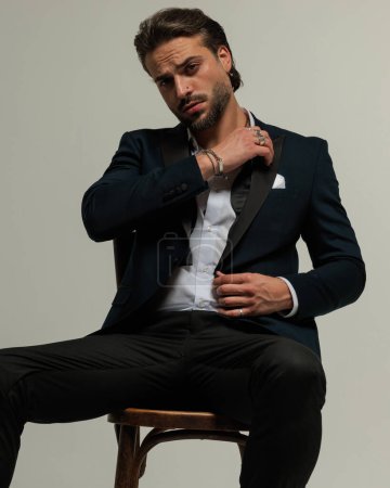 Téléchargez les photos : Élégant jeune homme d'affaires en tux assis et ajustant chemise col tout en étant assis devant fond gris - en image libre de droit