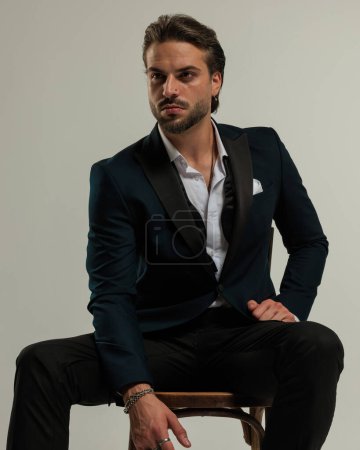 Téléchargez les photos : Homme d'affaires confiant en smoking avec nœud papillon défait regardant loin et assis sur une chaise en bois devant fond gris - en image libre de droit