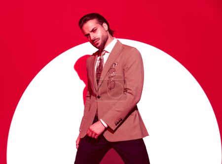 Foto de Elegante joven hombre de negocios mirando hacia adelante mientras sostiene los brazos en pose de moda frente al fondo rojo con foco - Imagen libre de derechos