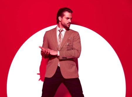 Téléchargez les photos : Portrait de sexy homme d'affaires barbu en costume regardant sur le côté et frotter les paumes devant fond rouge avec projecteur - en image libre de droit