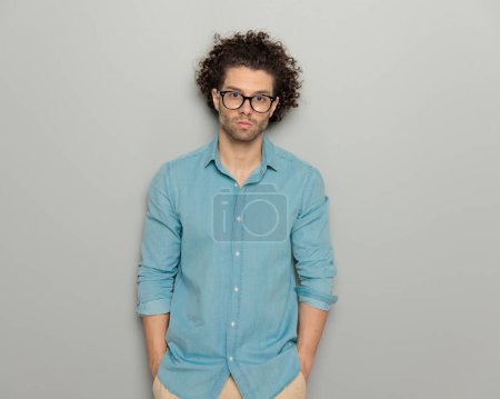 Téléchargez les photos : Homme sérieux occasionnel en denim bleu chemise avec des lunettes tenant la main et regardant vers l'avant en face de fond gris - en image libre de droit
