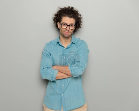 Photo for Portrait of casual curly hair man with glasses in blue denim shirt looking forward, smiling and crossing arms in front of grey background - Royalty Free Image