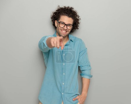 Téléchargez les photos : Portrait de jeune homme excité avec des lunettes souriant et pointant le doigt vers l'avant tout en tenant la main dans la poche devant fond gris - en image libre de droit