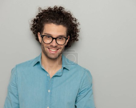 Téléchargez les photos : Portrait de jeune homme heureux avec des lunettes regardant vers l'avant et souriant devant fond gris - en image libre de droit