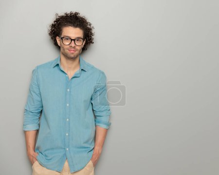 Téléchargez les photos : Homme cheveux bouclés avec des lunettes en denim chemise tenant la main dans les poches, souriant et regardant vers l'avant devant fond gris - en image libre de droit