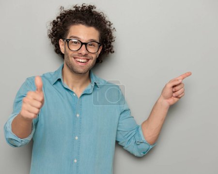 Photo for Handsome casual man with glasses pointing finger to side and making a thumbs up gesture in front of grey background - Royalty Free Image