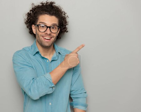 Photo for Attractive man with curly hair and glasses pointing fingers to side and showing, smiling and posing on grey background - Royalty Free Image