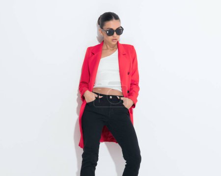 Téléchargez les photos : Sexy jeune femme en manteau rouge avec des lunettes de soleil tenant la main dans les poches et posant d'une manière fraîche sur fond blanc - en image libre de droit