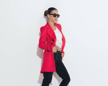 Téléchargez les photos : Femme de mode confiante avec des lunettes de soleil en manteau rouge regardant vers le côté et posant avec les mains dans les poches devant fond blanc - en image libre de droit