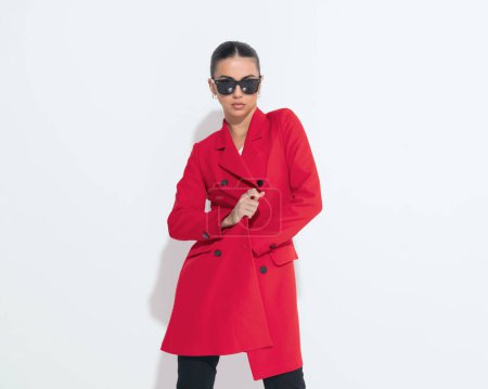 Foto de Mujer de moda cool con gafas de sol cerrando y ajustando el abrigo rojo mientras posa de una manera segura frente al fondo blanco - Imagen libre de derechos