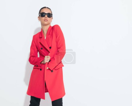 Téléchargez les photos : Jeune femme sexy avec des lunettes de soleil ajustant manteau rouge et posant devant fond blanc - en image libre de droit