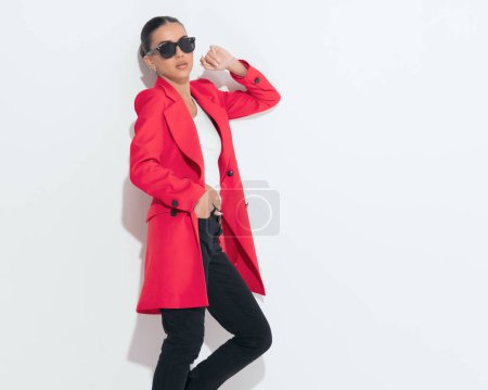 Téléchargez les photos : Attrayant jeune femme avec des lunettes de soleil tenant la main dans les poches et posant tout en posant sur un mur blanc en studio - en image libre de droit