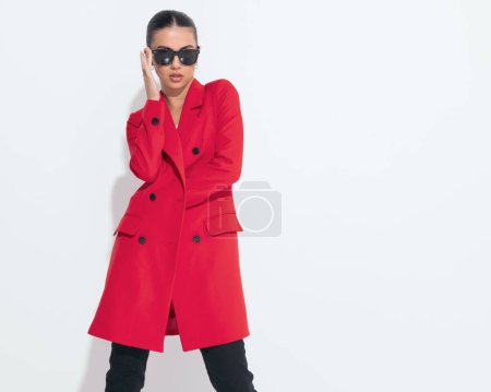 Téléchargez les photos : Portrait d'une jolie femme cool en manteau rouge fixant des lunettes de soleil et étant confiant tout en posant devant fond blanc - en image libre de droit