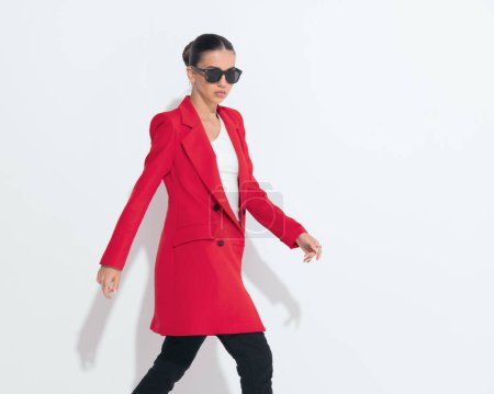 Téléchargez les photos : Cool femme élégante avec des lunettes de soleil en manteau rouge regardant loin et marchant devant fond blanc - en image libre de droit
